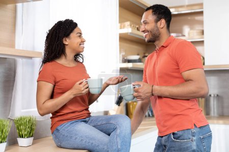 Cheerful millennial black couple in red t-shirts with cups of coffee enjoy communication in free time in kitchen interior, copy space. Drink favorite tea in morning, break and talk at home at weekend