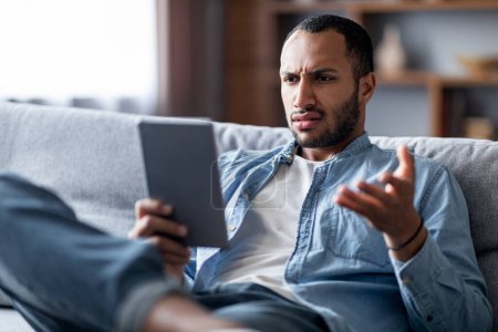Foto de Hombre negro joven estresado mirando la pantalla de la tableta digital y frunciendo el ceño mientras está sentado en el sofá en casa, hombre afroamericano confundido reaccionando emocionalmente a malas noticias o contenido en línea, primer plano - Imagen libre de derechos