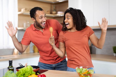 Téléchargez les photos : Heureux jeune homme noir et femme en t-shirts rouges s'amusent, chantant au microphone imaginaire à l'intérieur de la cuisine. Karaoké pour les superstars à la maison, couple appréciant la musique à temps libre et les émotions des gens - en image libre de droit