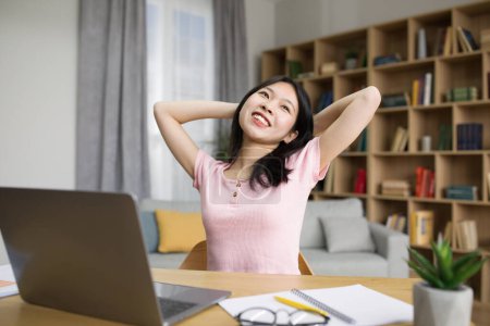 Téléchargez les photos : Étudiante asiatique détendue assise les mains derrière la tête près d'un ordinateur portable, ayant une pause après un cours en ligne à la maison, copiez l'espace. Enseignement à distance, concept de vidéoconférence pédagogique - en image libre de droit