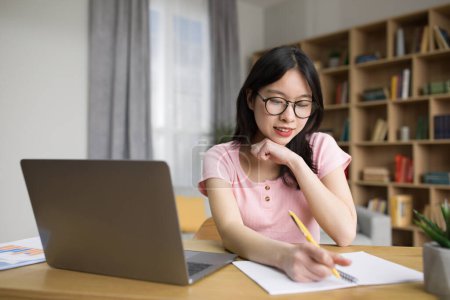 Photo for Positive korean woman having online lesson on laptop, writing in notebook while sitting at desk at home. Asian female student taking notes during webinar, studying remotely - Royalty Free Image