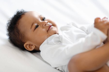 Foto de Primer plano Shot of Cute Little Black Baby Boy Of Girl acostado en la cama, Adorable pequeño niño afroamericano con traje de cuerpo Descansando en su espalda, Relajándose sobre sábanas blancas en el dormitorio en casa - Imagen libre de derechos