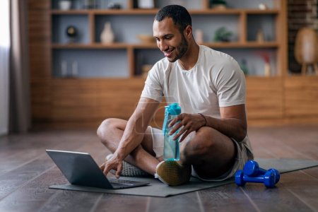Photo for Smiling Black Guy Opening Video Tutorials On Laptop Before Sport Training At Home, Happy Young African American Man Sitting On Fitness Mat In Living Room Interior And Using Computer, Copy space - Royalty Free Image