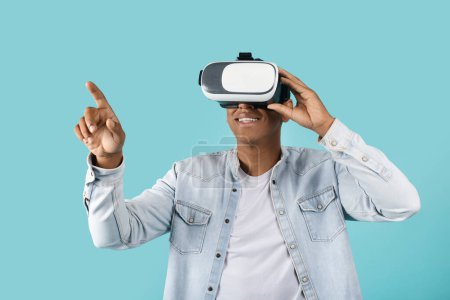 Foto de Sonriendo joven afroamericano masculino en gafas casuales y vr se divierte, disfrutar de la tecnología solo, el aire del tacto, aislado en el fondo azul, estudio. Mundo de realidad virtual y entretenimiento en tiempo libre - Imagen libre de derechos