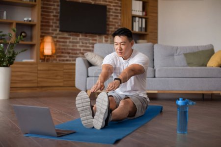 Photo for Home training concept. Active mature asian man stretching leg muscles in front of laptop, watching video tutorials online and doing pilates in living room, enjoying healthy lifestyle - Royalty Free Image
