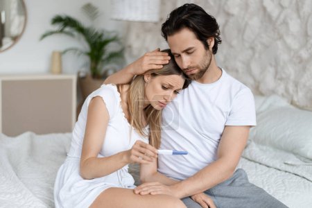Photo for Infertility problem. Caucasian couple sitting on bed with negative pregnancy test result, man embracing and calming his wife, free space - Royalty Free Image