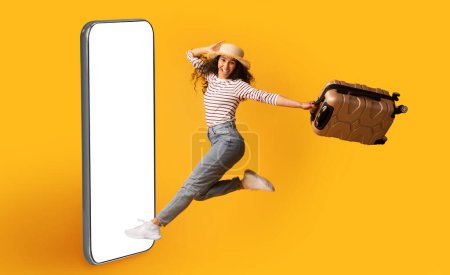 Photo for Emotional millennial brunette woman in casual comfy outfil tourist running towards big smartphone with white blank screen mockup for booking app, lady carrying luggage, yellow studio background - Royalty Free Image
