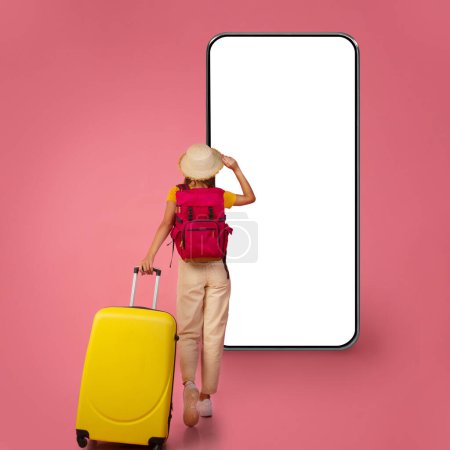 Téléchargez les photos : Vue arrière du sac à dos femme méconnaissable marchant avec une valise jaune sur fond de studio rose, touchant son chapeau d'été, regardant grand écran de maquette de téléphone portable, photo pleine longueur, espace de copie - en image libre de droit