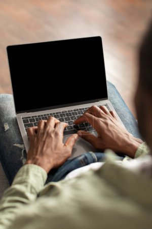 Reifer schwarzer Kerl, der auf der Computertastatur mit leerem Bildschirm tippt, empfiehlt Webseite im Wohnzimmerinnenraum, vertikal, unkenntlich. Arbeit und Geschäft aus der Ferne, freiberuflich, digitale Technologie und App