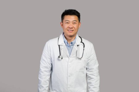 Téléchargez les photos : Souriant confiant bel homme chinois mature thérapeute en manteau blanc avec stéthoscope regardant la caméra, isolé sur fond gris, studio. Examen médical, soins de santé et traitement à l'hôpital - en image libre de droit
