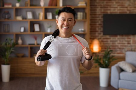 Photo for Active sporty lifestyle. Excited japanese middle aged man holding trx resistance bands hang around his neck, standing in living room and posing to camera - Royalty Free Image