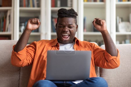 Online trading concept. Emotional happy millennial black man with dreadlocks in casual sitting on couch with modern laptop, looking at computer screen and gesturing, celebrating success, home interior