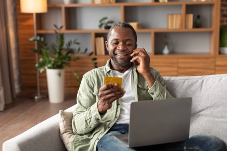 Foto de Feliz cliente hombre negro maduro feliz mirando la tarjeta de crédito y llamando por teléfono, ordena bienes con computadora en el interior de la sala de estar. Gestión financiera, compras en línea, venta y reembolso en casa - Imagen libre de derechos