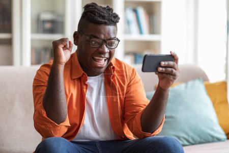Photo for Emotional chubby young african american guy sitting on couch in living room, holding smartphone, looking at phone screen, yelling, gesturing, celebrating success, trading on stocks and markets at home - Royalty Free Image