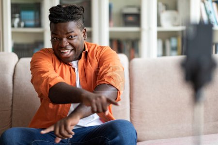 Téléchargez les photos : Célèbre influenceur afro-américain cool élégant chubby millénial gars enregistrement de contenu pour les médias sociaux, assis sur le canapé à la maison, regardant le téléphone portable sur le trépied, geste et sourire - en image libre de droit