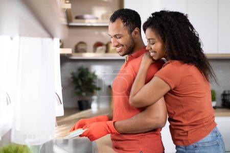 Photo for Household chores together at home. Glad millennial black wife hugging her husband in rubber gloves, guy washes dishes in kitchen interior, copy space. Love, romance, family relationship and lifestyle - Royalty Free Image