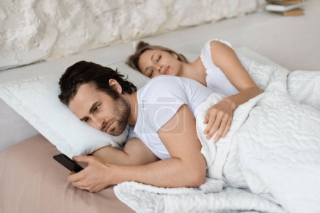 Photo for Angry man checking his sleeping girlfriends smartphone, lying in bed at home. Husband feeling jealous of her wifes infidelity, looking through her phone messages - Royalty Free Image