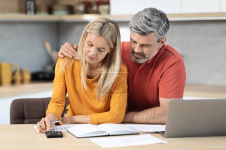 Photo for Middle aged spouses sitting at kitchen desk full of papers in front of computer, using calculator, mature man and woman counting incoming and outgoing money, planning family budget together - Royalty Free Image
