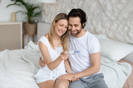 Photo for Happy husband and wife rejoicing positive pregnancy test, sitting on bed in bedroom interior, copy space. Good news, family planning, parenthood and expectation of child indoor - Royalty Free Image