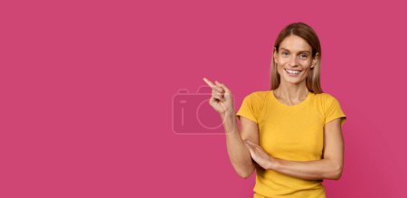 Foto de Sonriendo bastante mediana edad caucásica dama rubia delgada en camiseta amarilla señalando el dedo en el espacio libre, aislado en el fondo rosa, estudio. Asesoramiento y recomendación de profesionales, ofertas y anuncios - Imagen libre de derechos