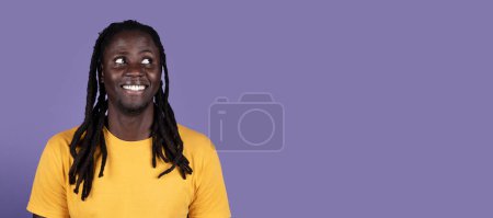 Foto de Sonriente chico negro joven guapo con rastas largas en camiseta amarilla mirando el espacio de copia en el fondo del estudio púrpura, comprobando gran oferta o oferta emocionante, web-banner, primer plano - Imagen libre de derechos