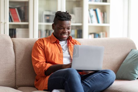 Téléchargez les photos : Carrière freelance. Heureux homme afro-américain en surpoids à l'aide d'un ordinateur portable assis sur le canapé à la maison, naviguant sur Internet et travaillant à distance en ligne. Travail à distance, Site Web, Espace de copie - en image libre de droit