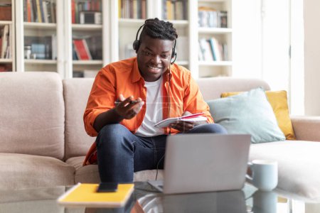 Photo for Cheerful overweight african american young man student sitting on couch at home, using headset and newest laptop, taking notes, attending webinar, copy space. Online education concept - Royalty Free Image
