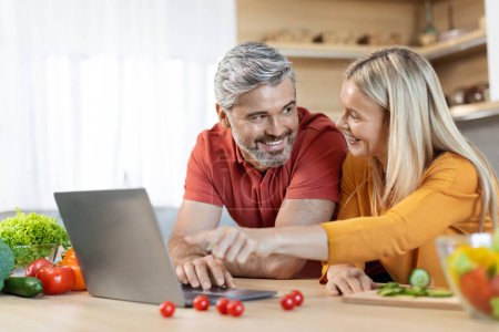 Photo for Cheerful attractive mature couple standing by kitchen desk full of fresh organic vegetables, using modern computer, pointing at laptop screen, looking for nice recipe on Internet, copy space - Royalty Free Image