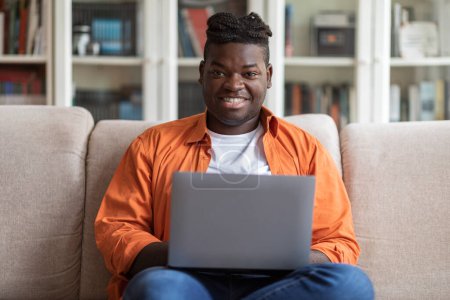 Photo for Remote job concept. Portrait of smiling happy young overweight african american guy in casual sitting on sofa, using modern laptop at home, data analyst typing on computer keyboard, copy space - Royalty Free Image