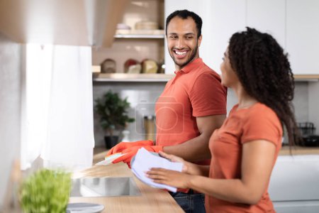 Photo for Cleaning and household chores together at home. Cheerful black millennial wife and husband in rubber gloves washes dishes in kitchen interior, free space. Love, romance, family relationship at weekend - Royalty Free Image