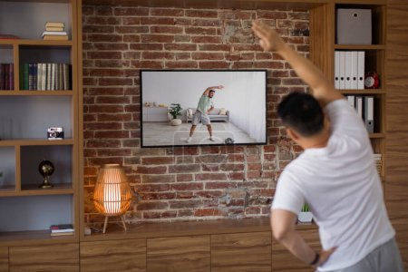 Photo for Sporty middle aged man working out at home, watching sport channel and fitness workout program on TV, back view. Healthy lifestyle concept - Royalty Free Image