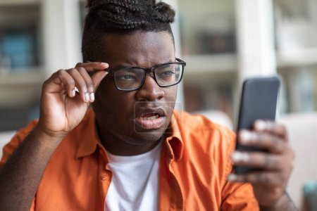 Foto de Confundido afroamericano joven se siente sorprendido recibir mal mensaje inesperado en el teléfono celular, frustrado gordito negro decepcionado por problemas con negocios tocar gafas, leer noticias, primer plano - Imagen libre de derechos
