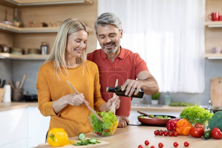 Photo for Good-looking caucasian middle aged spouses preparing healthy vegetable salad, happy beautiful wife mixing veggies in glass bowl, smiling husband adding olive oil rich in omega-3, kitchen interior - Royalty Free Image