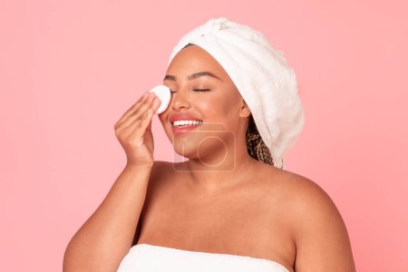 Photo for Attractive black plus size woman touching her face with cotton pad, removing makeup with cleansing milk, using toner or micellar water, pink studio background - Royalty Free Image