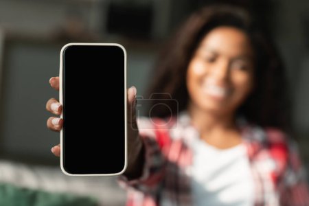Photo for Happy millennial black woman show smartphone with blank screen, recommending app, website in living room interior, blurred, close up. Online shopping, big sale and cashback, win, ad and offer at home - Royalty Free Image