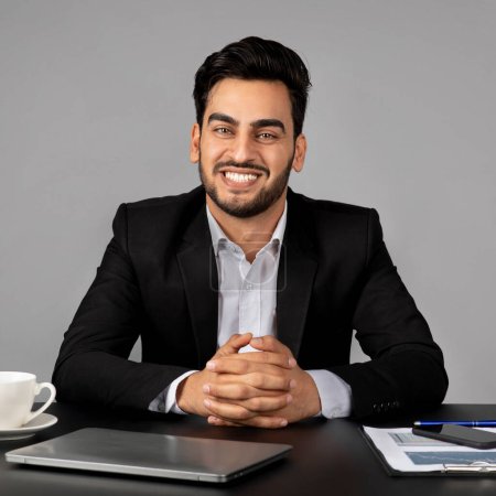 Foto de Retrato de joven empresario árabe guapo sentado en el escritorio sobre fondo gris y sonriendo en la cámara, empresario masculino de Oriente Medio feliz usando traje posando en el lugar de trabajo en la oficina, espacio de copia - Imagen libre de derechos