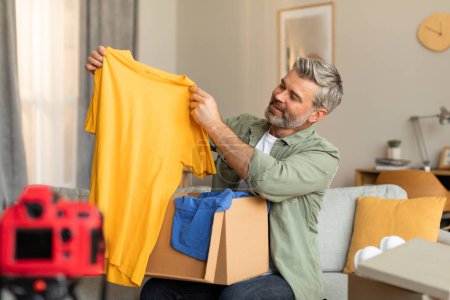 Photo for Cheerful elderly caucasian male blogger shoots vlog on camera with empty screen opens parcel shows clothes in living room interior. Fashion blog, review, sale, job and hobby, ad and offer at home - Royalty Free Image