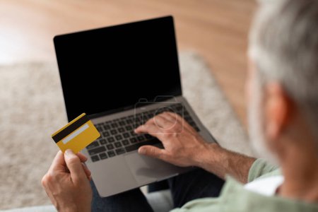 Photo for Cropped elderly caucasian man typing on computer with blank screen, using credit card, check finance in room interior. Modern technology, ad and offer, sale with cashback and online shopping at home - Royalty Free Image