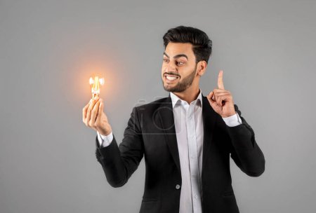 Foto de Ideas e innovaciones. Retrato de un joven empresario árabe sosteniendo una bombilla iluminada en la mano mientras está parado sobre un fondo gris en el estudio, un empresario masculino que tiene un momento inspirador - Imagen libre de derechos