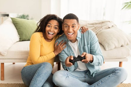Téléchargez les photos : Heureux jeune femme afro-américaine excitée et mari assis sur le sol avec joystick, jouer à un jeu en ligne dans le salon intérieur. Victoire, divertissement à la maison ensemble et plaisir avec la technologie moderne - en image libre de droit