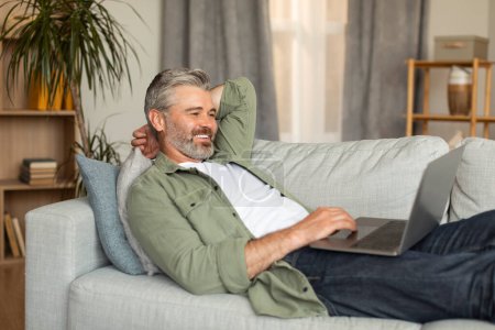 Téléchargez les photos : Homme caucasien âgé souriant tapant sur l'ordinateur, se reposant, couché sur le canapé et regardant la vidéo dans l'intérieur du salon. Technologie moderne pour l'appel vidéo, l'annonce et l'offre, le mode de vie et la détente à la maison - en image libre de droit