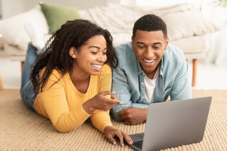 Photo for Happy young african american lady and man lie on floor, chatting on laptop, watch funny video, surfing in internet in living room interior. Modern technology, online game, video call at home together - Royalty Free Image