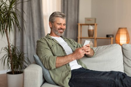 Photo for New normal for retired people. Smiling old caucasian male typing on smartphone uses new app for chat, video call in living room interior. Communication with modern technology, ad and offer at home - Royalty Free Image
