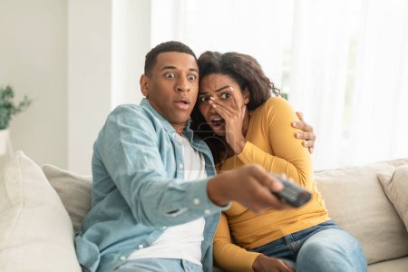 Photo for Scared shocked young african american woman and man with remote control switch channel on tv, watch horror film or bad news in living room interior. People emotion, fear, movie night at home together - Royalty Free Image