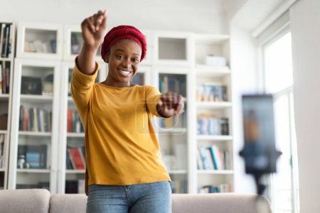 Téléchargez les photos : Célèbre influenceur afro-américain assez élégante dame millénaire en turban rouge enregistrement de contenu pour les adeptes dans les médias sociaux, la danse et le rire, en regardant le téléphone portable sur le trépied, copier l'espace - en image libre de droit