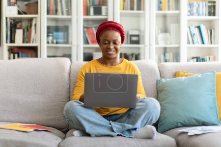 Téléchargez les photos : Jolie femme afro-américaine millénaire joyeuse avec turban rouge en tenue décontractée assise sur un canapé, tapant sur un clavier d'ordinateur portable et souriant, nomade numérique féminine travaillant de la maison, espace de copie - en image libre de droit