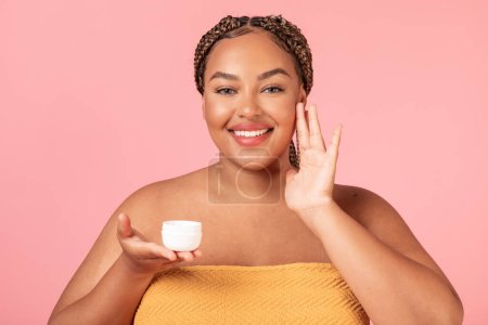 Photo for Bodycare concept. Happy black body positive lady applying beauty product on her face, using cream, standing wrapped in towel, posing over pink studio background - Royalty Free Image