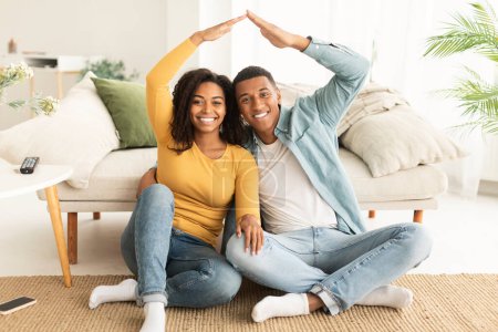 Téléchargez les photos : Souriant jeune femme afro-américaine et mari assis sur le sol faire toit avec leurs mains et regarder la caméra dans le salon intérieur. Crédit à l'immobilier, acheter sa propre maison, hypothèque et rêve de maison neuve - en image libre de droit