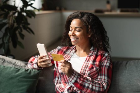 Photo for Glad smiling pretty millennial black lady checking bank account, ordering purchases with app, manage finance, buy in living room interior. Online shopping, big sale, pay money and cashback at home - Royalty Free Image