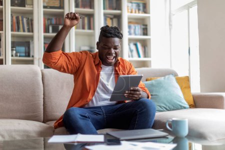 Téléchargez les photos : Millénaire africain dodu gars étudiant passer le test éducatif en ligne, jeune homme noir assis sur le canapé à la maison, tenant tablette numérique, lever la main, crier, célébrer le succès, copier l'espace - en image libre de droit